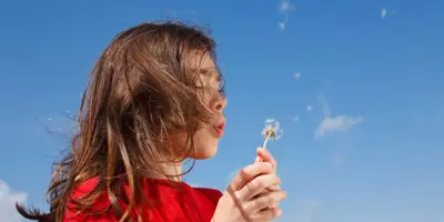 Una niña que podría necesitar algunos remedios caseros para la fiebre del heno mientras sopla un diente de león con un cielo azul de fondo